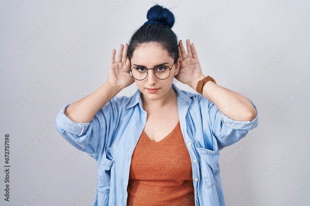 Young modern girl with blue hair standing over white background trying to hear both hands on ear gesture, curious for gossip. hearing problem, deaf Young modern girl with blue hair standing over white background trying to hear both hands on ear gesture, curious for gossip. hearing problem, deaf
