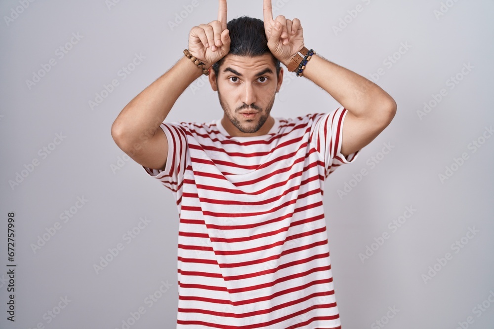 Hispanic man with long hair standing over isolated background doing funny gesture with finger over head as bull horns Hispanic man with long hair standing over isolated background doing funny gesture with finger over head as bull horns