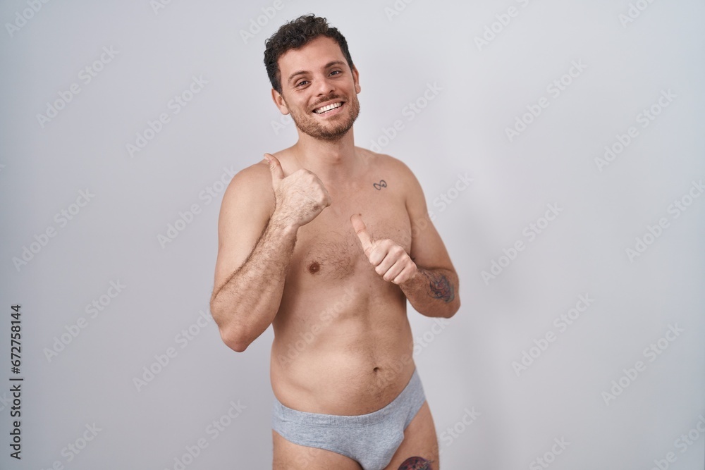 Young hispanic man standing shirtless wearing underware pointing to the back behind with hand and thumbs up, smiling confident