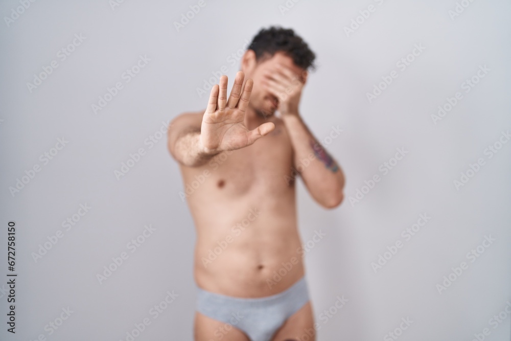 Fototapeta premium Young hispanic man standing shirtless wearing underware covering eyes with hands and doing stop gesture with sad and fear expression. embarrassed and negative concept.