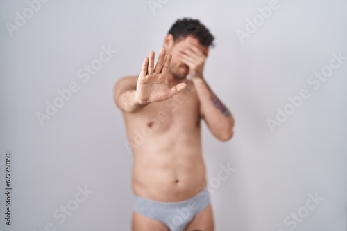 Young hispanic man standing shirtless wearing underware covering eyes with hands and doing stop gesture with sad and fear expression. embarrassed and negative concept.