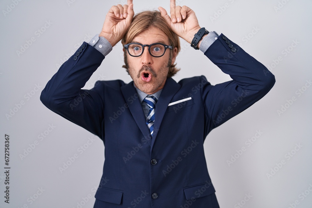 Caucasian man with mustache wearing business clothes doing funny gesture with finger over head as bull horns Caucasian man with mustache wearing business clothes doing funny gesture with finger over head as bull horns