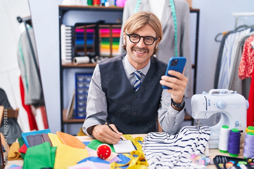 Young blond man tailor using smartphone writing on notebook at clothing factory Young blond man tailor using smartphone writing on notebook at clothing factory