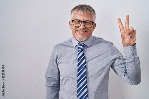 Hispanic business man with grey hair wearing glasses smiling looking to the camera showing fingers doing victory sign. number two.