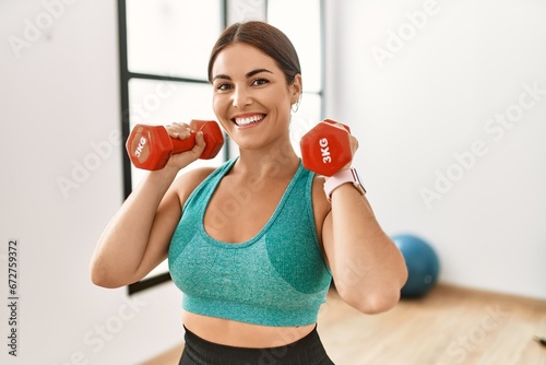 Young beautiful hispanic woman smiling confident using dumbbells training at sport center
