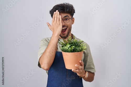 Arab man with beard holding green plant pot covering one eye with hand, confident smile on face and surprise emotion.