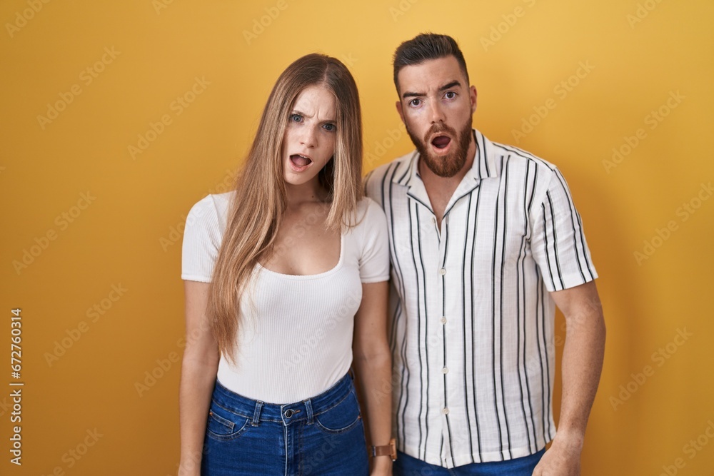 Young couple standing over yellow background in shock face, looking skeptical and sarcastic, surprised with open mouth Young couple standing over yellow background in shock face, looking skeptical and sarcastic, surprised with open mouth