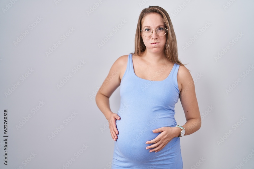 Young pregnant woman standing over white background puffing cheeks with funny face. mouth inflated with air, crazy expression.