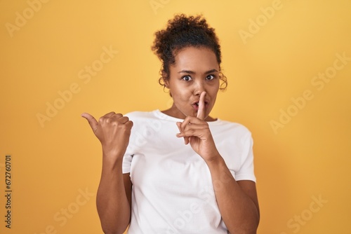 Young hispanic woman with curly hair standing over yellow background asking to be quiet with finger on lips pointing with hand to the side. silence and secret concept.