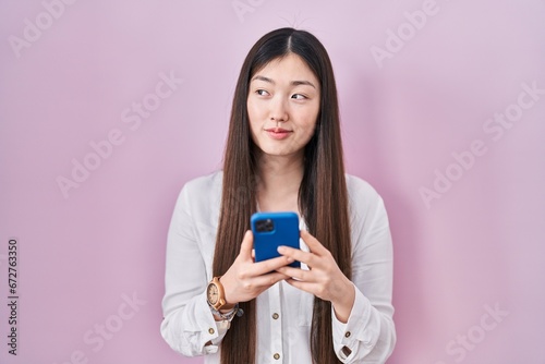 Chinese young woman using smartphone typing message smiling looking to the side and staring away thinking.