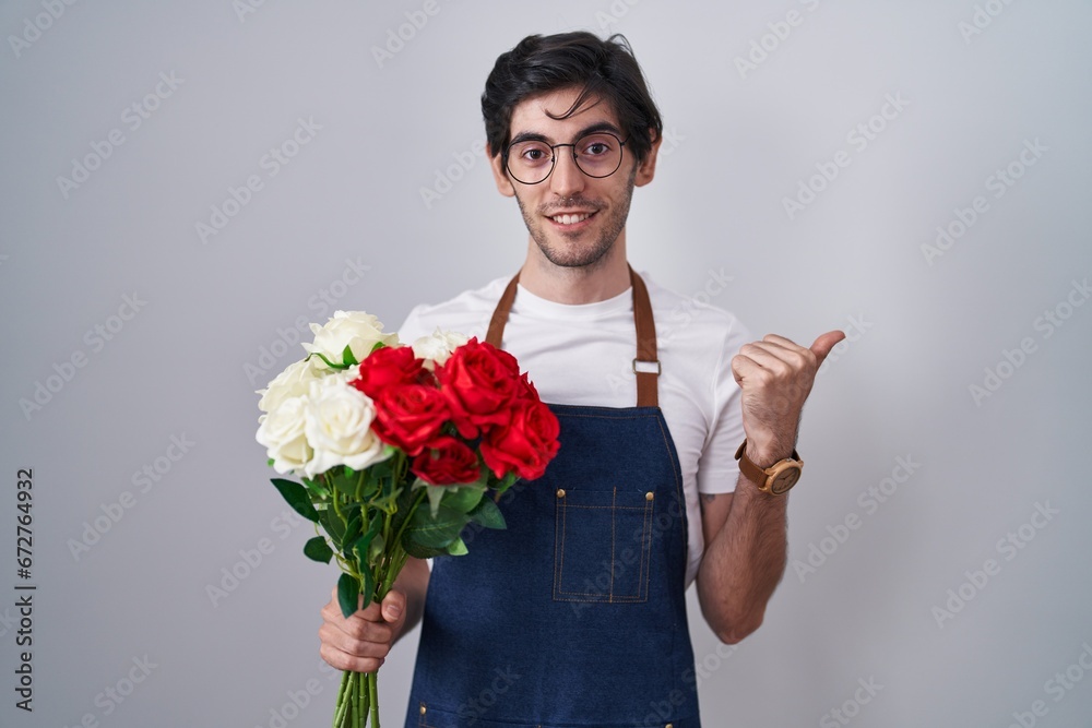 Young hispanic man holding bouquet of white and red roses pointing to the back behind with hand and thumbs up, smiling confident Young hispanic man holding bouquet of white and red roses pointing to the back behind with hand and thumbs up, smiling confident