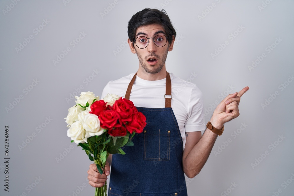 Young hispanic man holding bouquet of white and red roses surprised pointing with finger to the side, open mouth amazed expression. Young hispanic man holding bouquet of white and red roses surprised pointing with finger to the side, open mouth amazed expression.