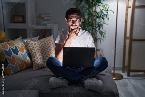 Young hispanic man using laptop at home at night thinking worried about a question, concerned and nervous with hand on chin
