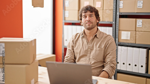 Young hispanic man ecommerce business worker sitting on table with serious face at office