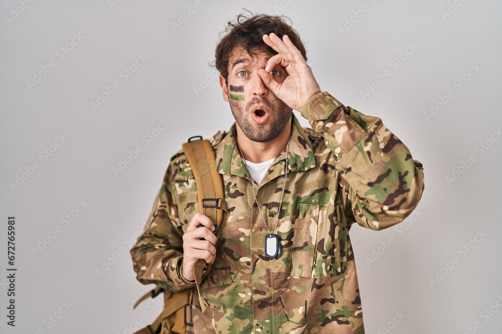 Hispanic young man wearing camouflage army uniform doing ok gesture shocked with surprised face, eye looking through fingers. unbelieving expression.