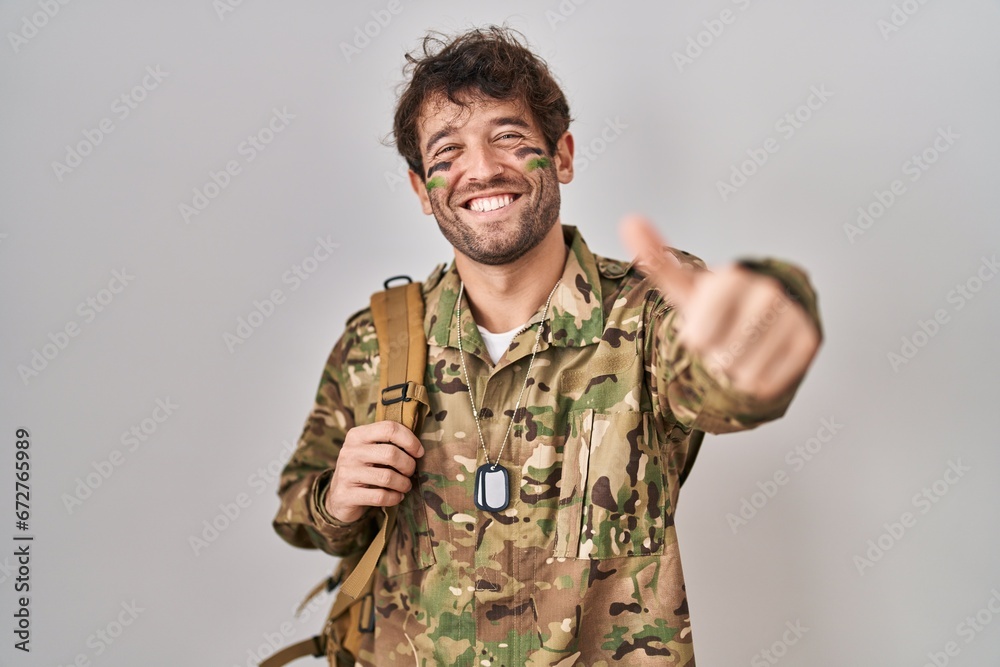 Hispanic young man wearing camouflage army uniform approving doing positive gesture with hand, thumbs up smiling and happy for success. winner gesture. Hispanic young man wearing camouflage army uniform approving doing positive gesture with hand, thumbs up smiling and happy for success. winner gesture.