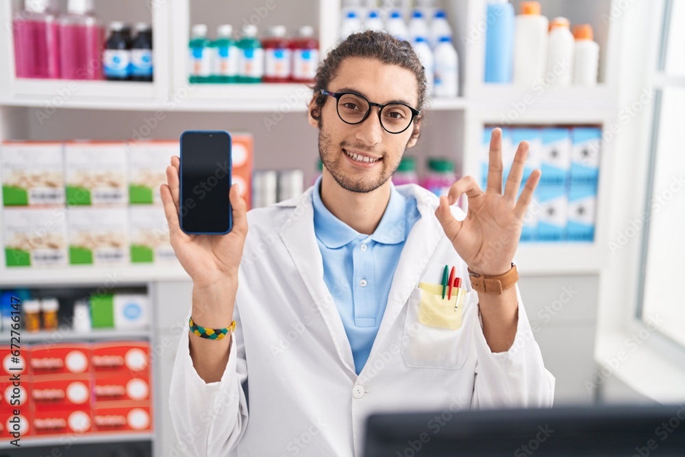 Young hispanic man working at pharmacy drugstore showing smartphone screen doing ok sign with fingers, smiling friendly gesturing excellent symbol