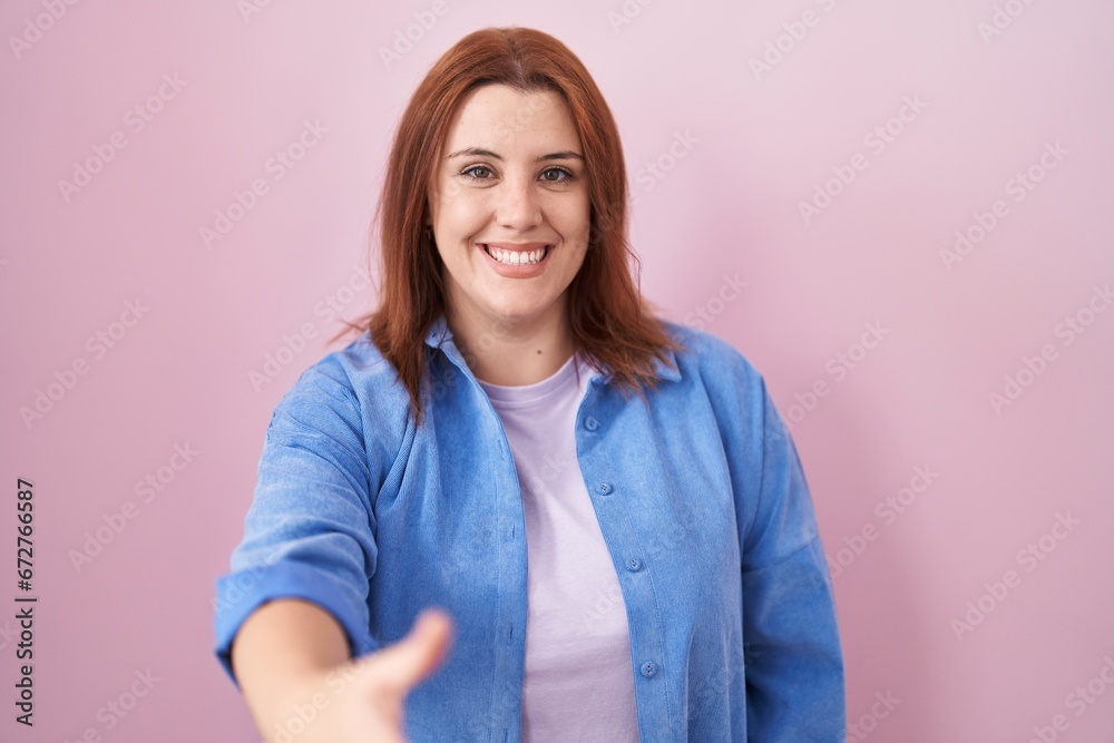 Fototapeta premium Young hispanic woman with red hair standing over pink background smiling friendly offering handshake as greeting and welcoming. successful business.