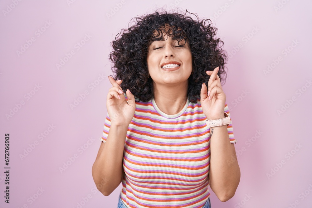 Young middle east woman standing over pink background gesturing finger crossed smiling with hope and eyes closed. luck and superstitious concept. Young middle east woman standing over pink background gesturing finger crossed smiling with hope and eyes closed. luck and superstitious concept.