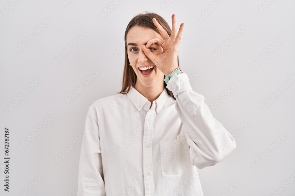 Young caucasian woman standing over isolated background doing ok gesture with hand smiling, eye looking through fingers with happy face. Young caucasian woman standing over isolated background doing ok gesture with hand smiling, eye looking through fingers with happy face.