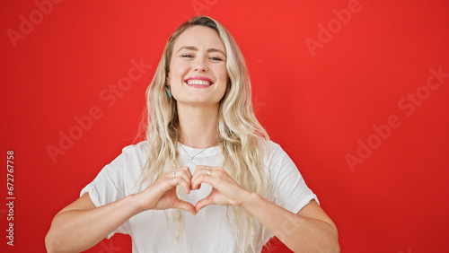 Young blonde woman smiling confident doing heart gesture with hands over isolated red background photo