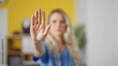Young blonde woman doctor doing stop sign with hand at clinic