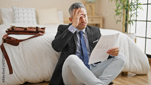 Young hispanic man business worker reading document stressed sitting on floor at hotel room