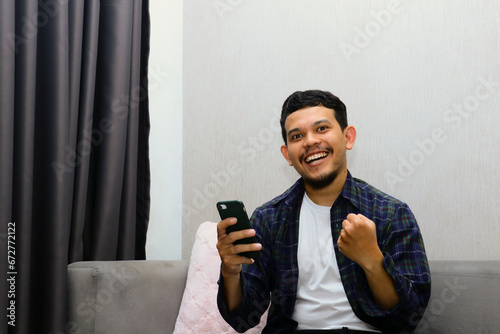 Happy young man sitting on a gray sofa and holding a mobile phone. Guy browsing internet, surfing web, using app, smiling expression photo