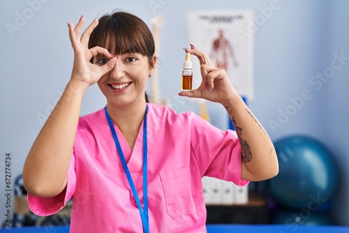 Young brunette woman holding cbd oil at physiotherapy clinic smiling happy doing ok sign with hand on eye looking through fingers