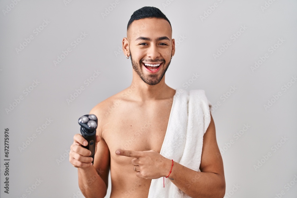 Young hispanic man holding electric razor smiling happy pointing with hand and finger - obrazy, fototapety, plakaty 
