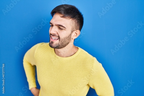 Hispanic man standing over blue background suffering of backache, touching back with hand, muscular pain