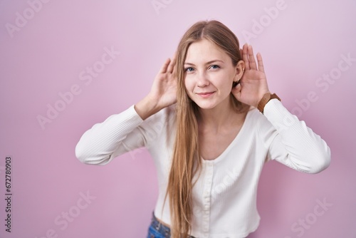 Young caucasian woman standing over pink background trying to hear both hands on ear gesture, curious for gossip. hearing problem, deaf