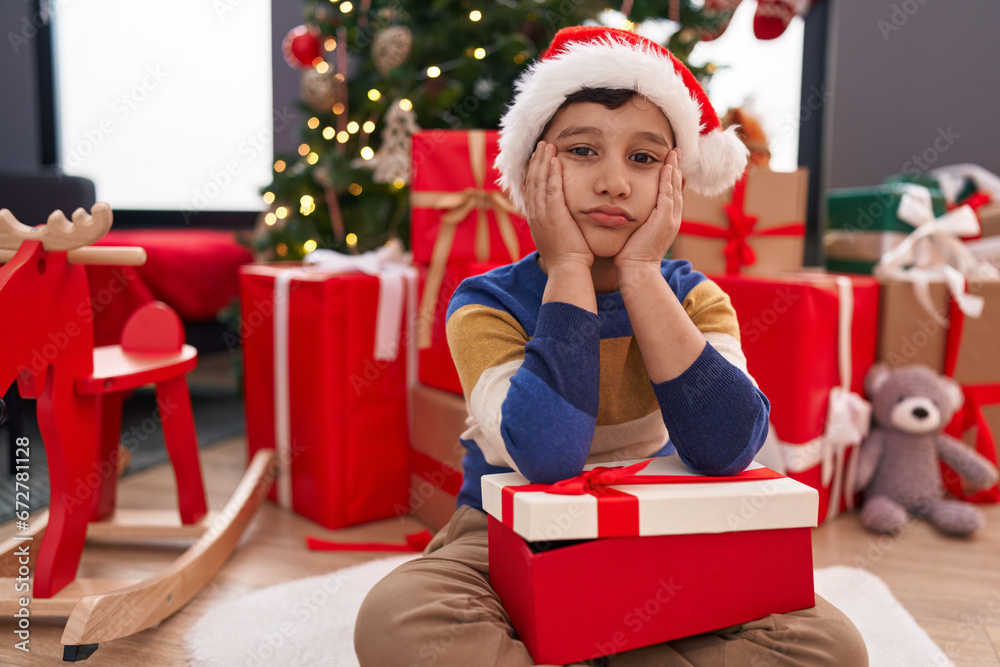 Adorable hispanic boy holding christmas gift sitting on floor with unhappy expression at home Adorable hispanic boy holding christmas gift sitting on floor with unhappy expression at home