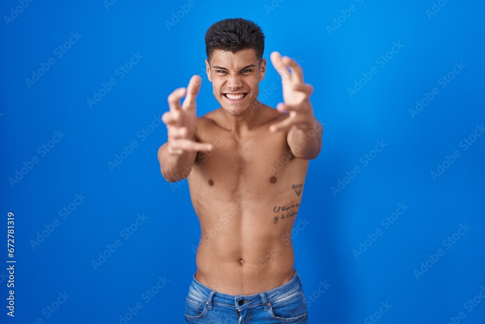 Young hispanic man standing shirtless over blue background shouting frustrated with rage, hands trying to strangle, yelling mad