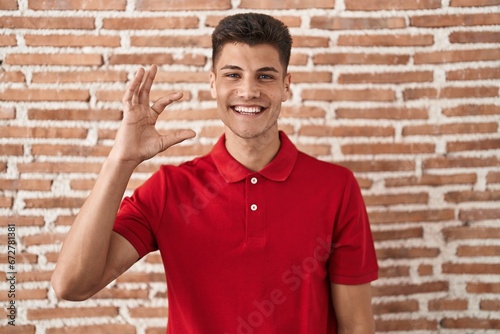 Young hispanic man standing over bricks wall smiling and confident gesturing with hand doing small size sign with fingers looking and the camera. measure concept.