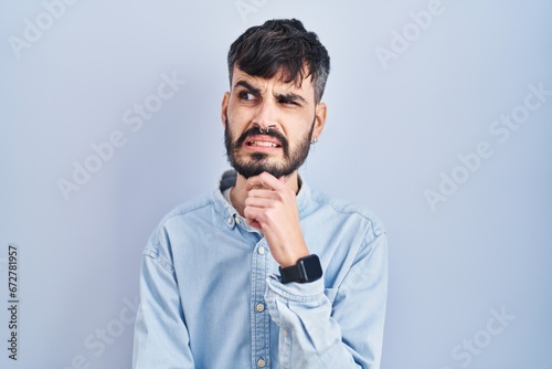 Young hispanic man with beard standing over blue background thinking worried about a question, concerned and nervous with hand on chin