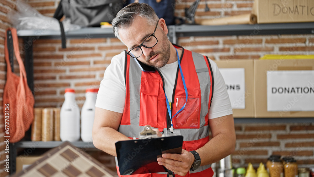 Grey-haired man volunteer writing on checklist talking on smartphone at charity center