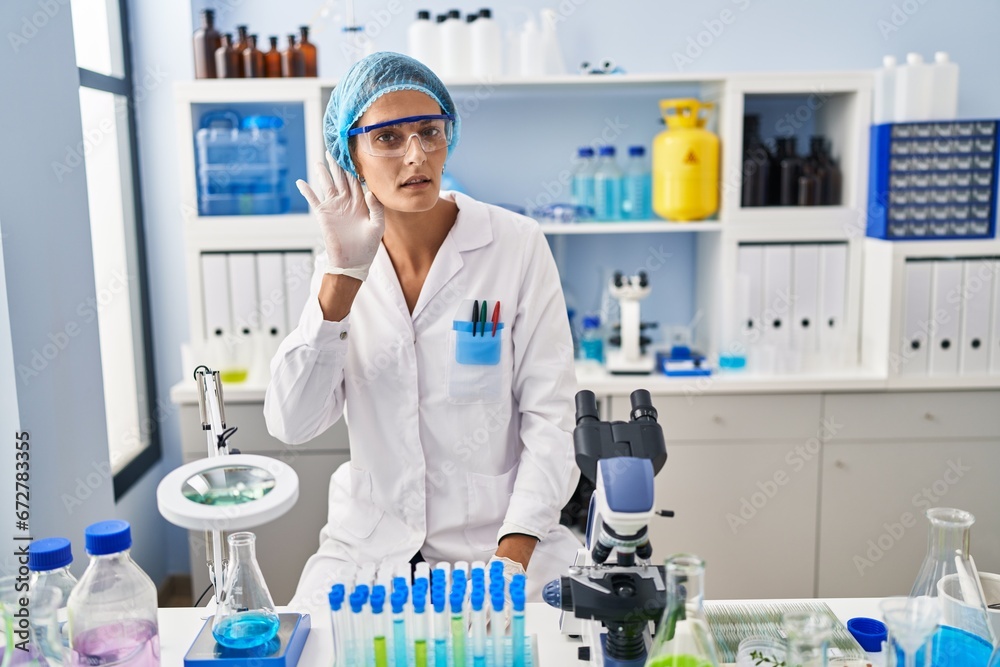 Brunette woman working at scientist laboratory smiling with hand over ear listening an hearing to rumor or gossip. deafness concept. Brunette woman working at scientist laboratory smiling with hand over ear listening an hearing to rumor or gossip. deafness concept.
