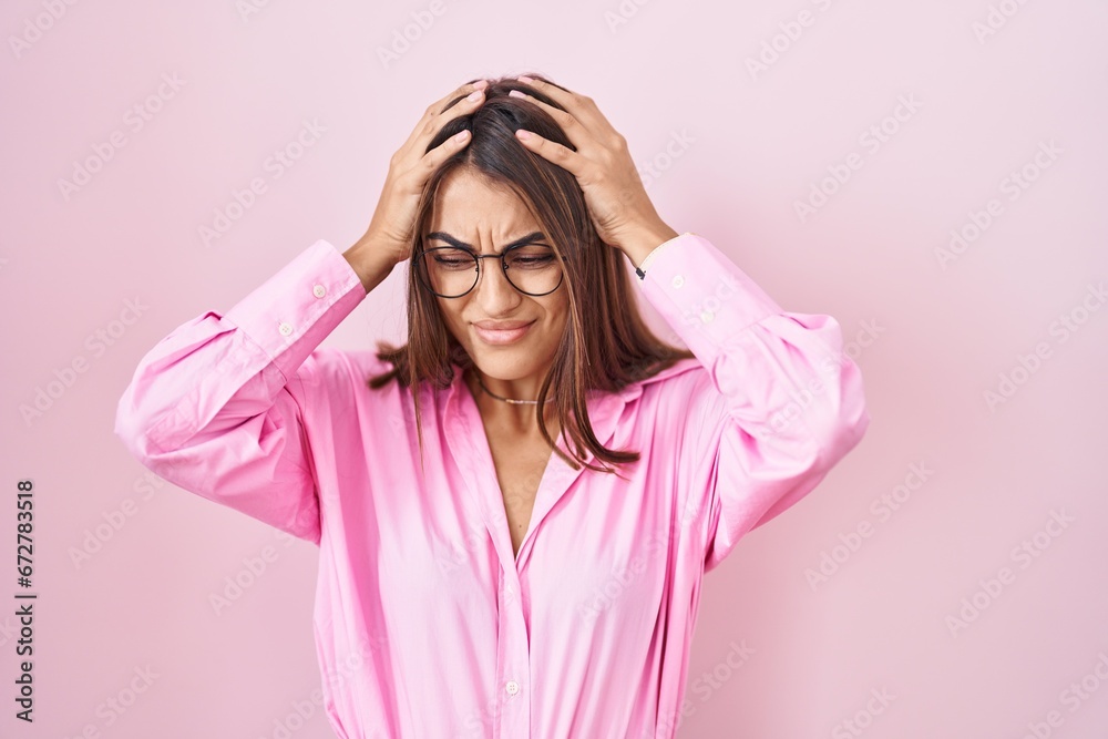 Young hispanic woman wearing glasses standing over pink background suffering from headache desperate and stressed because pain and migraine. hands on head. Young hispanic woman wearing glasses standing over pink background suffering from headache desperate and stressed because pain and migraine. hands on head.