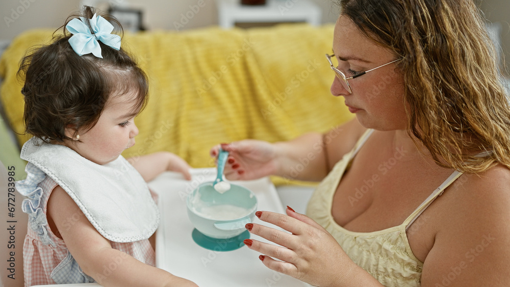 Mother and daughter together, loving family lifestyle, casually sitting at home during baby's lunch time, holding spoon and bib, concentrating on eating in the living room interior. Mother and daughter together, loving family lifestyle, casually sitting at home during baby's lunch time, holding spoon and bib, concentrating on eating in the living room interior.