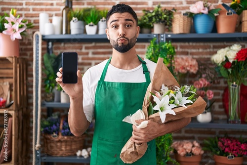 Hispanic young man working at florist shop showing smartphone screen looking at the camera blowing a kiss being lovely and sexy. love expression.