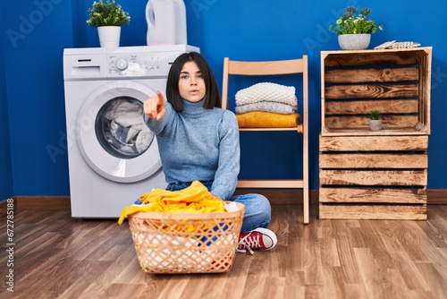 Young hispanic woman at laundry room pointing with finger to the camera and to you, confident gesture looking serious