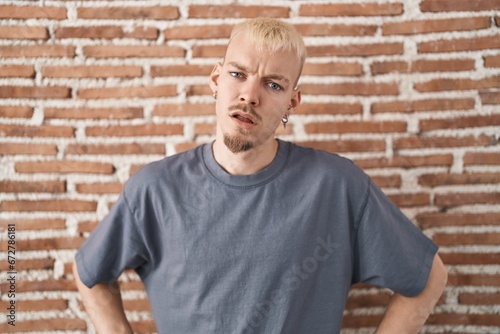 Young caucasian man standing over bricks wall in shock face, looking skeptical and sarcastic, surprised with open mouth