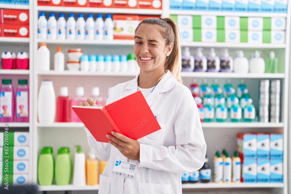 Young blonde woman working at pharmacy drugstore holding notebook winking looking at the camera with sexy expression, cheerful and happy face.