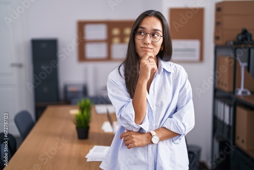 Young hispanic woman at the office with hand on chin thinking about question, pensive expression. smiling with thoughtful face. doubt concept. photo