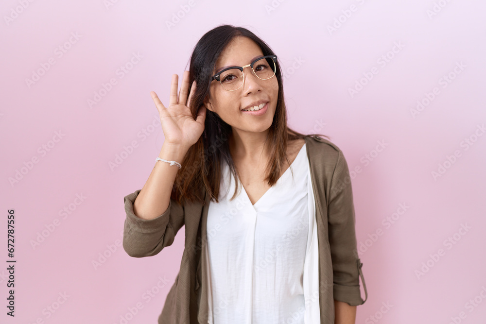 Middle age chinese woman wearing glasses over pink background smiling with hand over ear listening an hearing to rumor or gossip. deafness concept. Middle age chinese woman wearing glasses over pink background smiling with hand over ear listening an hearing to rumor or gossip. deafness concept.