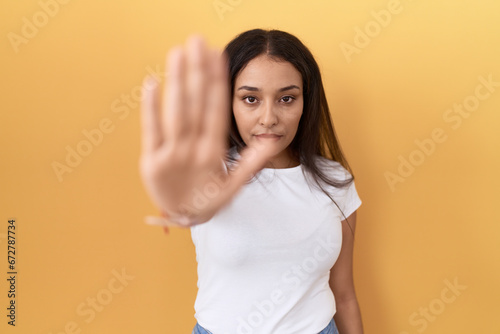 Young arab woman wearing casual white t shirt over yellow background doing stop sing with palm of the hand. warning expression with negative and serious gesture on the face.