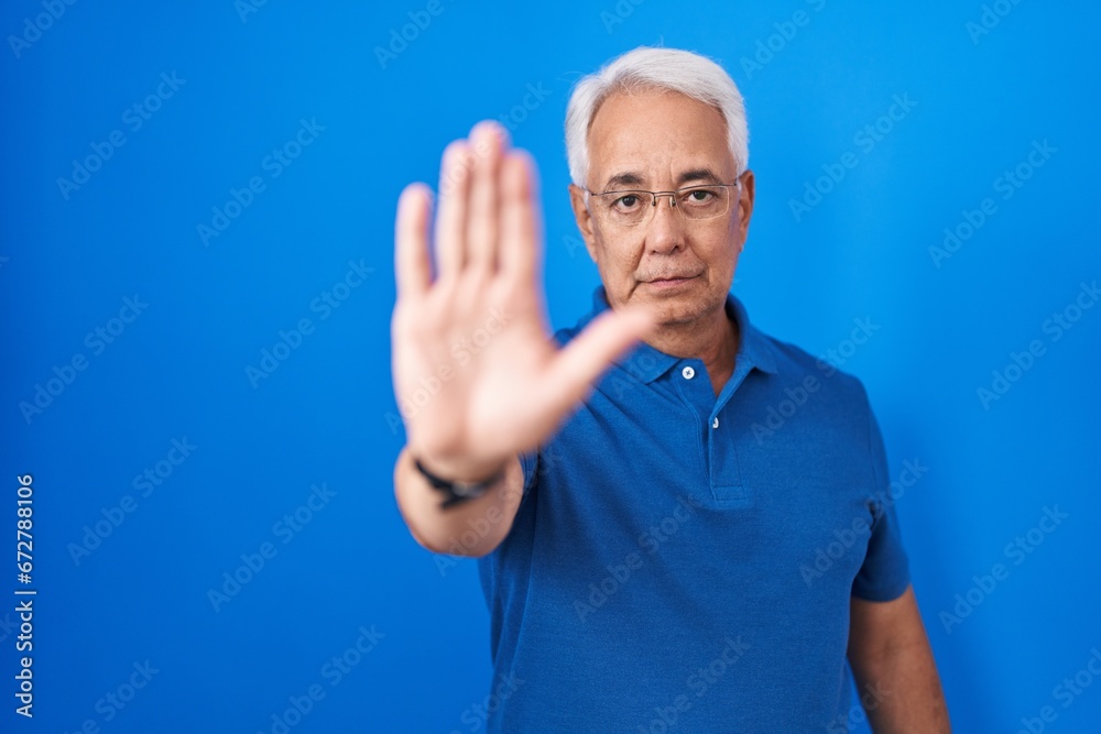 Middle age man with grey hair standing over blue background doing stop sing with palm of the hand. warning expression with negative and serious gesture on the face. Middle age man with grey hair standing over blue background doing stop sing with palm of the hand. warning expression with negative and serious gesture on the face.