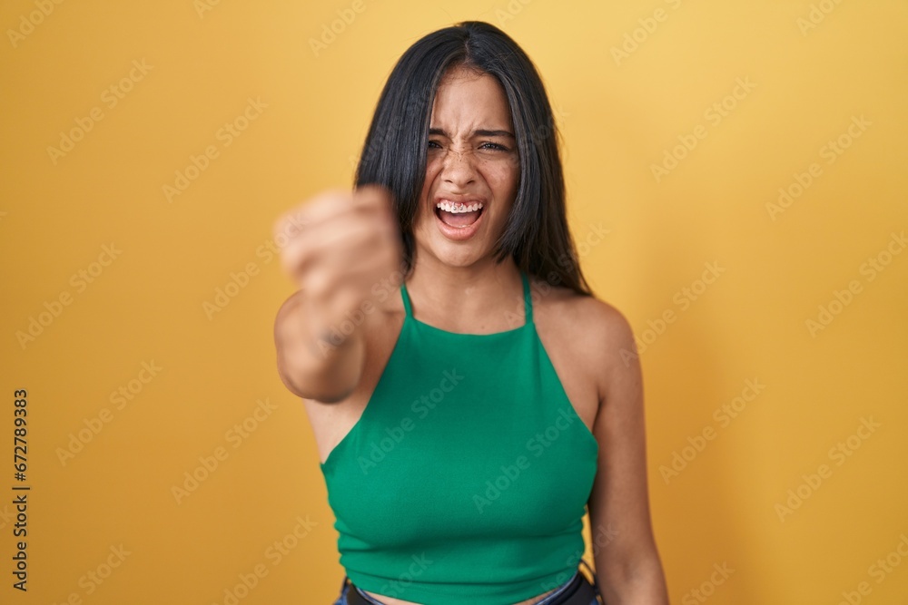 Brunette woman standing over yellow background angry and mad raising fist frustrated and furious while shouting with anger. rage and aggressive concept.