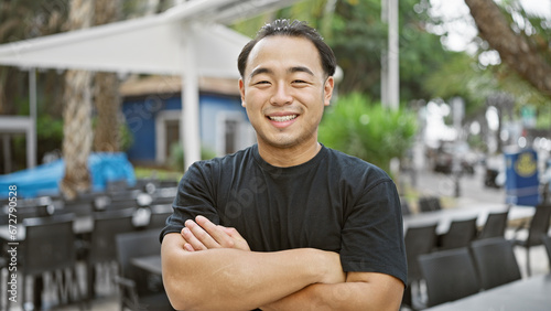 Cheerful young chinese man, arms crossed, confidently standing on coffee shop terrace, letting his joy radiate through his relaxed smiling expression while enjoying urban outdoors.